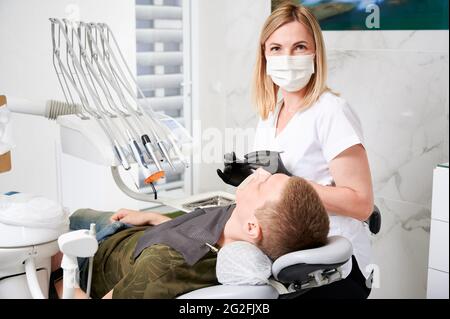 Instantané horizontal d'une jolie, jeune femme dentiste, portant uniforme, masque facial, gants en caoutchouc, assis à son lieu de travail dans un cabinet dentaire bien équipé. Patient masculin lors de la visite du dentiste Banque D'Images