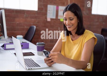 Femme d'affaires caucasienne souriante assise au bureau avec un ordinateur portable, à l'aide d'un smartphone Banque D'Images