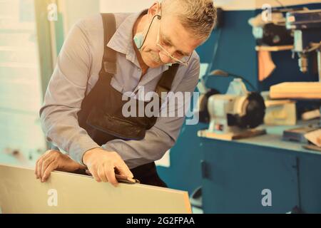 Portrait d'un menuisier âgé ou d'un charpentier en combinaison travaillant avec des planches en bois dans un atelier de menuisier. Sciage de bois. Banque D'Images