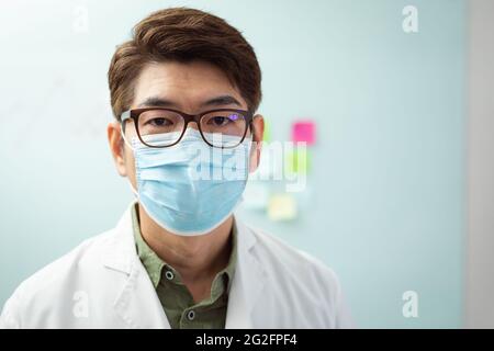 Portrait d'un médecin asiatique de sexe masculin portant un masque, assis dans le bureau de l'hôpital et regardant un appareil photo Banque D'Images