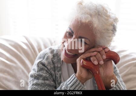 Femme âgée de race mixte assise sur un canapé tenant une canne à pied et souriante Banque D'Images