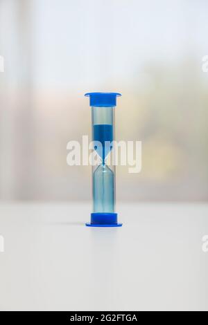 Un sablier bleu sur une surface blanche. La partie supérieure est pleine car le compte à rebours a commencé. En arrière-plan il y a une fenêtre à travers laquelle beaucoup o Banque D'Images