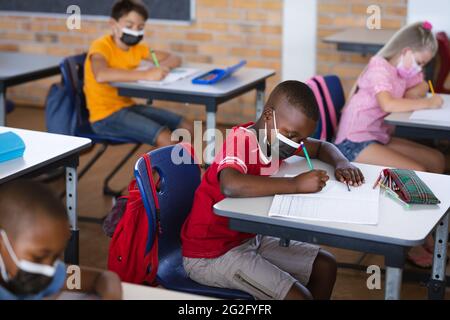 Un garçon afro-américain portant un masque facial étudiant assis sur son bureau en classe à l'école Banque D'Images