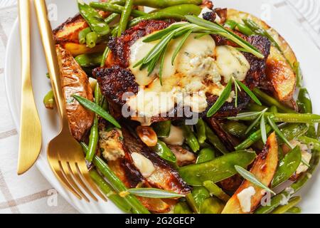 steak de porc recouvert de fromage bleu fondu et de romarin sur une bande de pois mange-tout grillés, de quartiers de pommes de terre, de haricots verts et d'ail Banque D'Images