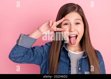 Photo portrait de petite fille montrant V-signe geste souriant montrant la langue isolée sur fond rose pastel Banque D'Images