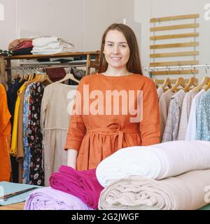 Positive femme propriétaire d'atelier posant près de rouleaux de tissu et de robes sur le fond Banque D'Images