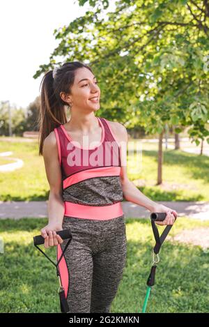 vue verticale d'une femme de race blanche s'exerçant avec des bandes de résistance. Elle sourit et porte des vêtements de sport roses et gris Banque D'Images