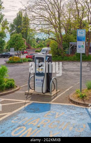 Point de charge pour les véhicules électriques situés dans un parking. Une aire de stationnement bleue spécifique est prévue en face. Les arbres et les arbustes abondent. Banque D'Images