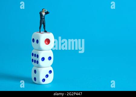 Homme d'affaires utilisant des jumelles se tenant au-dessus de trois piles de dés. Photo miniature de jouets de personnes minuscules. Isolé sur fond bleu. Banque D'Images