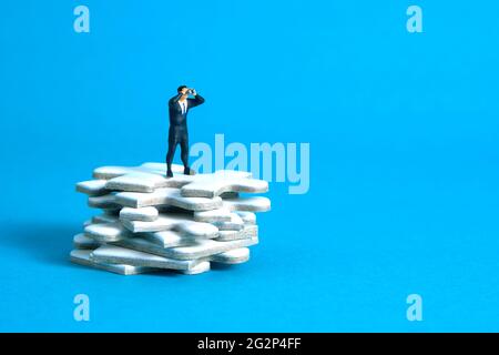 Les hommes d'affaires se tenant au-dessus de la pile de pièces de puzzle utilisant des jumelles au-dessus de la table en bois. Photo miniature de jouets de personnes minuscules. Isolé sur bac bleu Banque D'Images