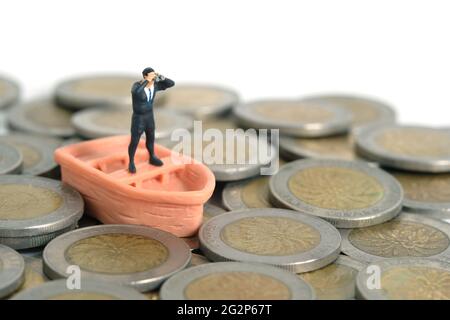 Homme d'affaires utilisant des jumelles se tenant au-dessus du bateau et pile de monnaie. Photo miniature de jouets de personnes minuscules. Isolé sur fond blanc Banque D'Images