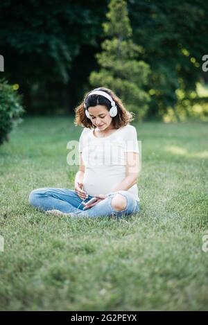 enceinte dans un casque à l'extérieur. Une jeune femme heureuse assise sur un tapis et écoutant de la musique sur un casque depuis un smartphone au parc. Détente, détente a Banque D'Images