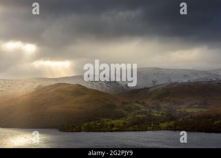 Des rayons de lumière spectaculaires traversent des nuages sombres au-dessus des montagnes Cumbriennes enneigées entourant Ullswater dans le Lake District.