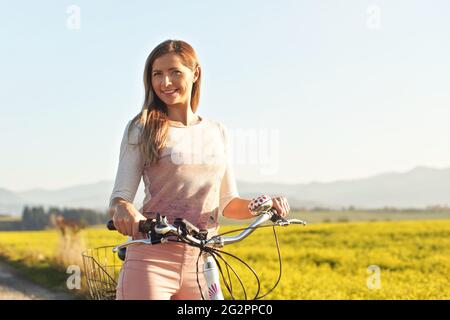 Une jeune femme se tenant à côté de son vélo sur une route de campagne poussiéreuse, un fort contre-jour de soleil de l'après-midi en arrière-plan brille sur le champ de fleurs jaunes Banque D'Images