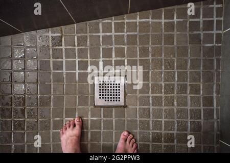 Vue sur la grille de drainage de la douche de la chambre d'hôtel, carrelage humide au sol, pieds d'homme visibles dans la partie inférieure Banque D'Images