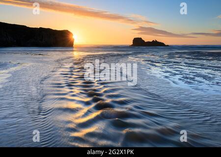 Le sable se déforme et une rivière mène l'œil du spectateur vers Chapel Rock sur la plage Perranporth de Cornwall. L'image a été capturée peu avant le coucher du soleil. Banque D'Images