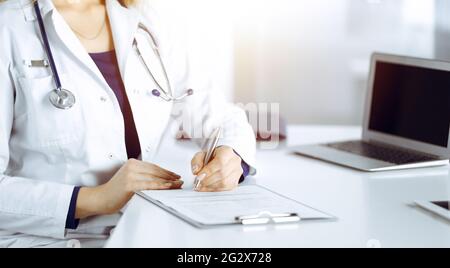 Une femme-médecin inconnue prescrit des médicaments à son patient, à l'aide d'un presse-papiers, tout en étant assise au bureau dans son cabinet ensoleillé. Femme Banque D'Images