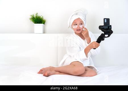 Une jeune fille blogueuse est assise sur un lit dans un peignoir blanc avec une serviette sur sa tête, des taches sur le visage sur son visage, enregistre une vidéo vlog sur une caméra à la maison Banque D'Images