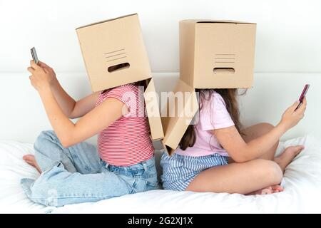 Deux adolescentes avec des boîtes sur leur tête sont assises et regardent les smartphones sur le lit à la maison. Le visage et les yeux sont recouverts d'une boîte. Le c Banque D'Images