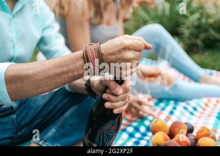 Gros plan les mains d'un homme ouvrant un vin mousseux tout en étant assis sur une couverture avec sa femme célébrant la vie, en appréciant l'un l'autre Banque D'Images