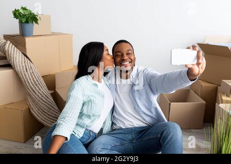 Amoureux des conjoints noirs se déplaçant à leur propre maison, prenant le selfie parmi les boîtes en carton, femme embrassant mari dans la joue Banque D'Images