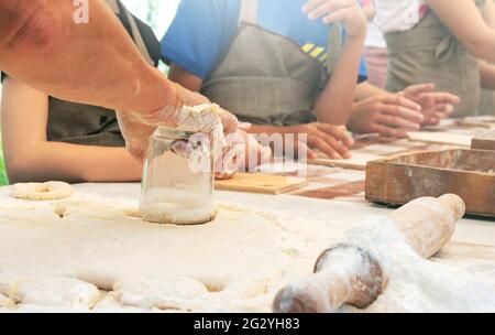 La femme coupe les cercles de la pâte. Le cuisinier enseigne aux enfants à faire des beignets sucrés. Classe de maître dans la cuisson. Cuisiner à la maison. Banque D'Images