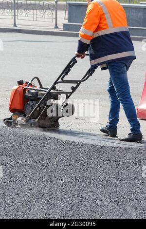 Un travailleur routier compacte de l'asphalte frais avec une plaque vibrante à essence par temps ensoleillé. Banque D'Images