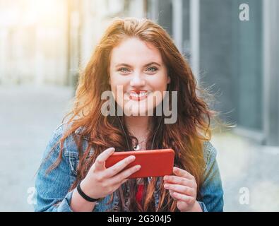 Image de concept des personnes modernes avec des appareils technologiques. Portrait de cheveux longs à boucles rouges jeune fille caucasienne marchant dans la rue et parcourant l'inter Banque D'Images