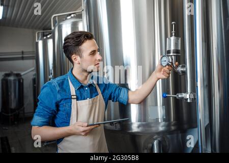 Un travailleur de brasserie vérifie le processus de fermentation dans la cuve d'acier et regarde le bloc-notes de l'usine Banque D'Images