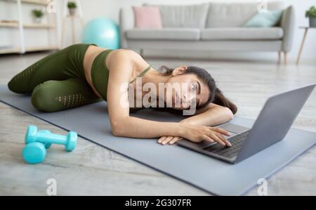 Sports en ligne. Une jeune femme indienne fatiguée, allongé sur un tapis de yoga près d'un ordinateur portable, n'ayant aucune force pour l'entraînement domestique Banque D'Images