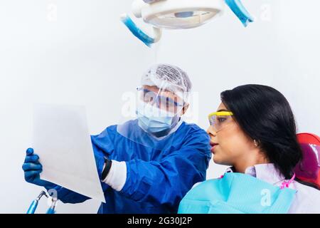 Un dentiste masculin adulte portant des gants et un costume de biosécurité montrant une radiographie orale à une patiente Banque D'Images
