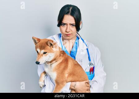 Belle femme vétérinaire hispanique tenant chien sceptique et nerveux, frowning bouleversé à cause du problème. Personne négative. Banque D'Images