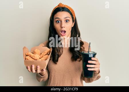 Jeune femme hispanique mangeant des ailes de poulet et buvant du soda peur et choqué par l'expression surprise et stupéfiée, la peur et le visage excité. Banque D'Images