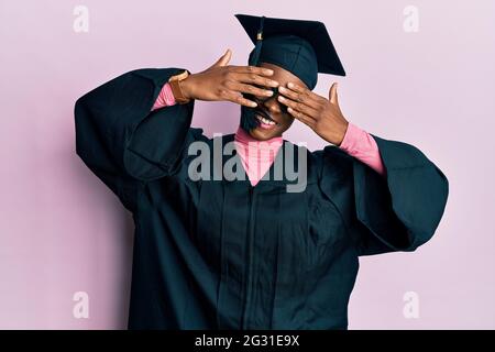 Jeune fille afro-américaine portant un chapeau de remise de diplômes et une robe de cérémonie couvrant les yeux avec les mains souriantes et drôles. Concept aveugle. Banque D'Images