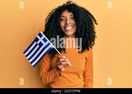 Femme afro-américaine aux cheveux afro tenant le drapeau grec à l'air positif et heureux debout et souriant avec un sourire confiant montrant des dents Banque D'Images