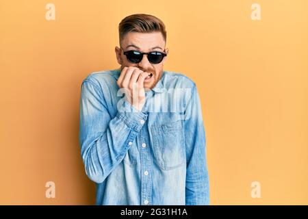 Jeune homme à tête rouge portant des lunettes de soleil stylées qui ont l'air stressé et nerveux avec les mains sur la bouche piquant les ongles. Problème d'anxiété. Banque D'Images