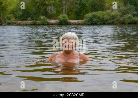 Portrait en gros plan d'une femme âgée souriante qui pose dans l'eau de la rivière en un jour d'automne nuageux. Banque D'Images