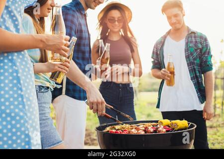 l'homme frite de la viande et des légumes sur le gril à l'air libre, tourne des saucisses avec des pinces en gros plan, des épis de maïs, des piments et des ailes de poulet, des frises Banque D'Images