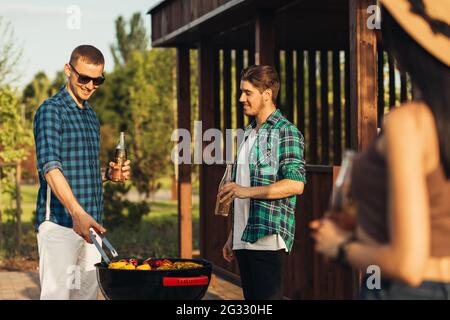 Deux jeunes hommes avec des boissons à la bière, faisant le barbecue dans la nature, les gens à la mode de préparation de la viande, le mode de vie des jeunes, la nourriture, l'amitié et le concept d'été Banque D'Images