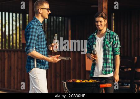 Deux jeunes hommes avec des boissons à la bière, faisant le barbecue dans la nature, les gens à la mode de préparation de la viande, le mode de vie des jeunes, la nourriture, l'amitié et le concept d'été Banque D'Images