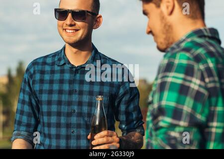 Deux jeunes hommes avec des boissons à la bière, faisant le barbecue dans la nature, les gens à la mode de préparation de la viande, le mode de vie des jeunes, la nourriture, l'amitié et le concept d'été Banque D'Images