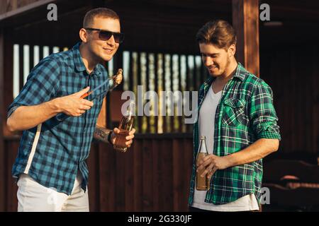 Deux jeunes hommes avec des boissons à la bière, faisant le barbecue dans la nature, les gens à la mode de préparation de la viande, le mode de vie des jeunes, la nourriture, l'amitié et le concept d'été Banque D'Images