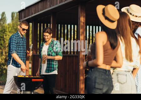 Deux jeunes hommes avec des boissons à la bière, faisant le barbecue dans la nature, les gens à la mode de préparation de la viande, le mode de vie des jeunes, la nourriture, l'amitié et le concept d'été Banque D'Images