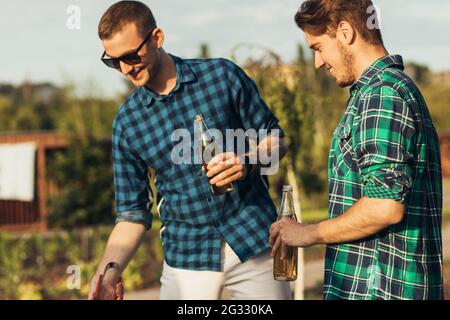 Deux jeunes hommes avec des boissons à la bière, faisant le barbecue dans la nature, les gens à la mode de préparation de la viande, le mode de vie des jeunes, la nourriture, l'amitié et le concept d'été Banque D'Images