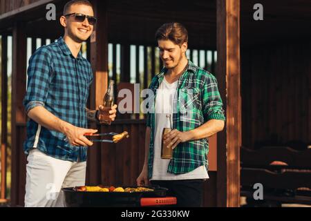 Deux jeunes hommes avec des boissons à la bière, faisant le barbecue dans la nature, les gens à la mode de préparation de la viande, le mode de vie des jeunes, la nourriture, l'amitié et le concept d'été Banque D'Images