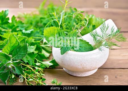 Feuilles et branches de menthe, d'aneth, d'estragon, de thym, de céleri et de persil dans un mortier en pierre avec un pilon et sur une table contre le fond de bois b Banque D'Images