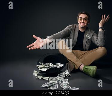 Joyeux sourire homme d'âge moyen en veste, jeans et chaussures est assis sur le sol par chapeau à l'envers plein de dollars de trésorerie gesturant Banque D'Images