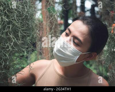 Belle femme asiatique portant un masque médical blanc propre et une chemise sans manches beige et regardant la plante suspendue à l'envers dans le jardin vert. H Banque D'Images