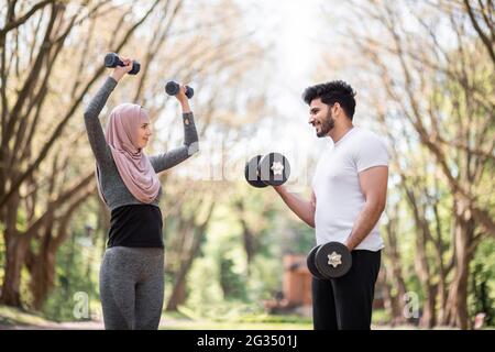 Vue latérale d'un couple musulman en bonne santé dans les vêtements de sport ayant l'entraînement à l'extérieur pendant l'heure du matin. Les jeunes hommes et femmes s'entrainement de muscles avec des haltères. Banque D'Images