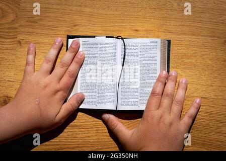 Les mains des enfants tiennent une bible penchée sur une table en bois. Petit livre Sainte Bible.Kid lecture de la sainte bible,closeup. Banque D'Images